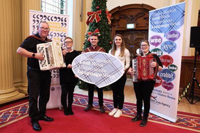 Line up of five people with two holding accordians with banners and a prop promoting Ulster-Scots Language Week