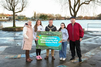 Belfast Lord Mayor pictured with community reps beside Waterworks lake with sign saying 'Reconnecting Waterworks Park and Alexandra Park development proposals public consultation'