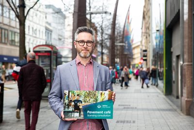 Councillor McKeown stands on a pavement in city centre with a poster promoting council's pavement cafe scheme