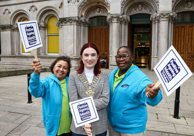 Three women stand outside a grand building holding signs that say Positive Ageing Month