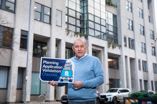 Councillor Arder Carson standing in front of an office building holding a sign promoting council's Planning Application Validation Checklist