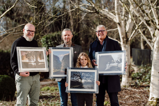 Four people, winners of the Belfast Parks Photographic Competition, hold winning framed photographs with trees behind them