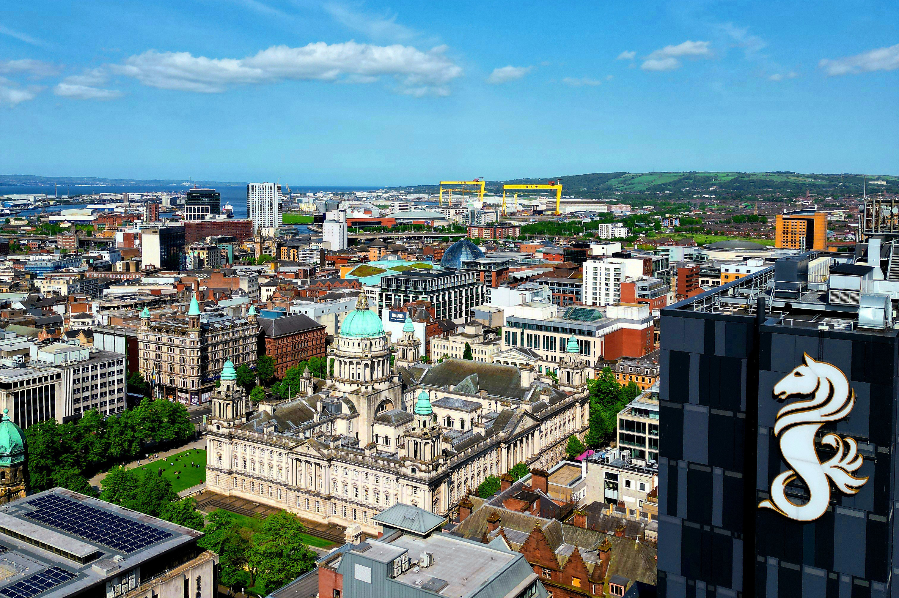 Aerial shot of Belfast city centre featuring Grand Central Hotel and City Hall against blue sky.