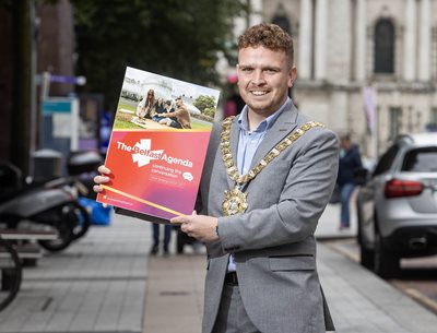 Belfast Lord Mayor Councillor Ryan Murphy holding a copy of the updated Belfast Agenda document with Donegall Place and City Hall in background