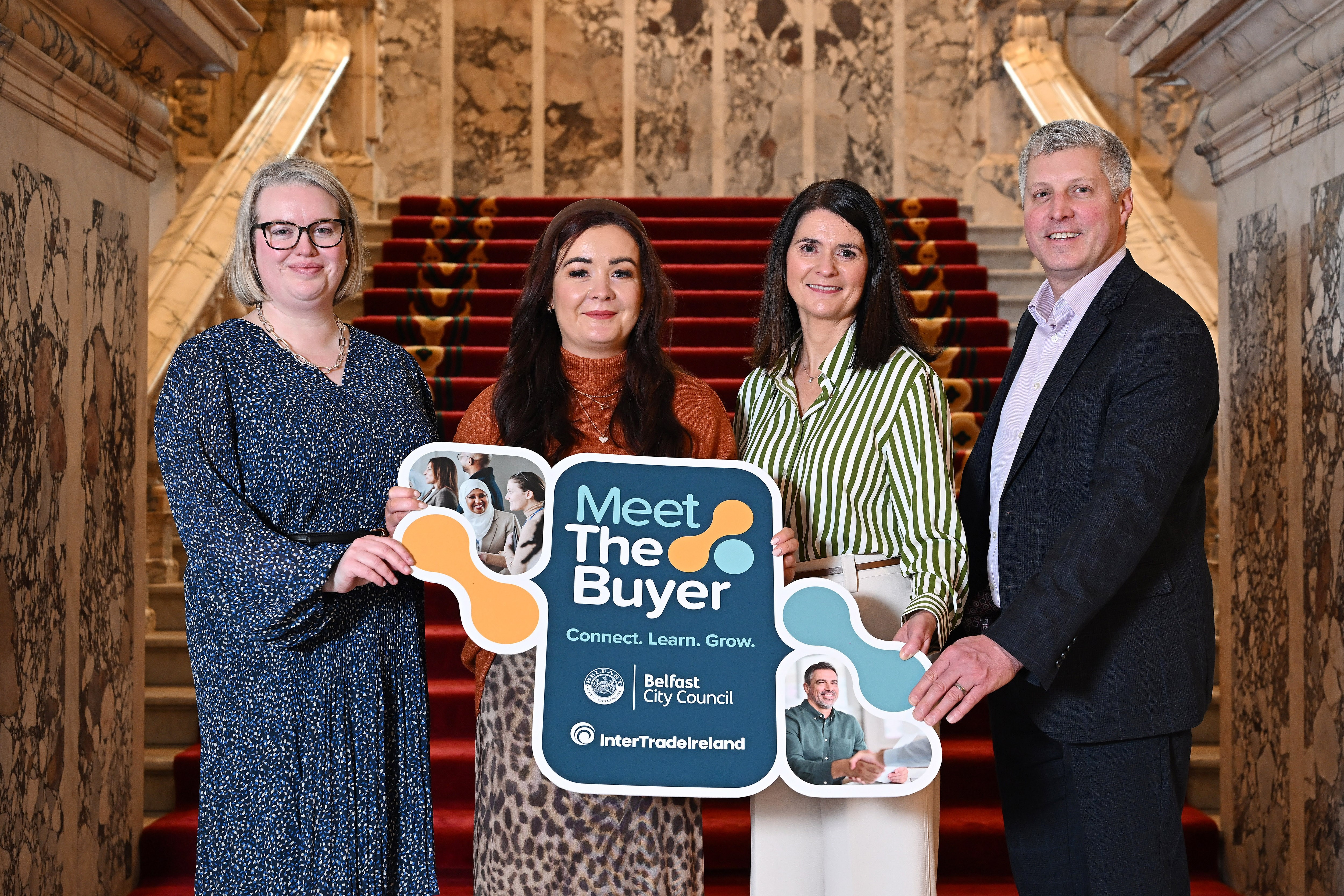 Four people including Councillor Natasha Brennan holding a 'Meet the Buyer' promotional artwork for the launch of the event taking place in  Belfast City Hall in March 2026