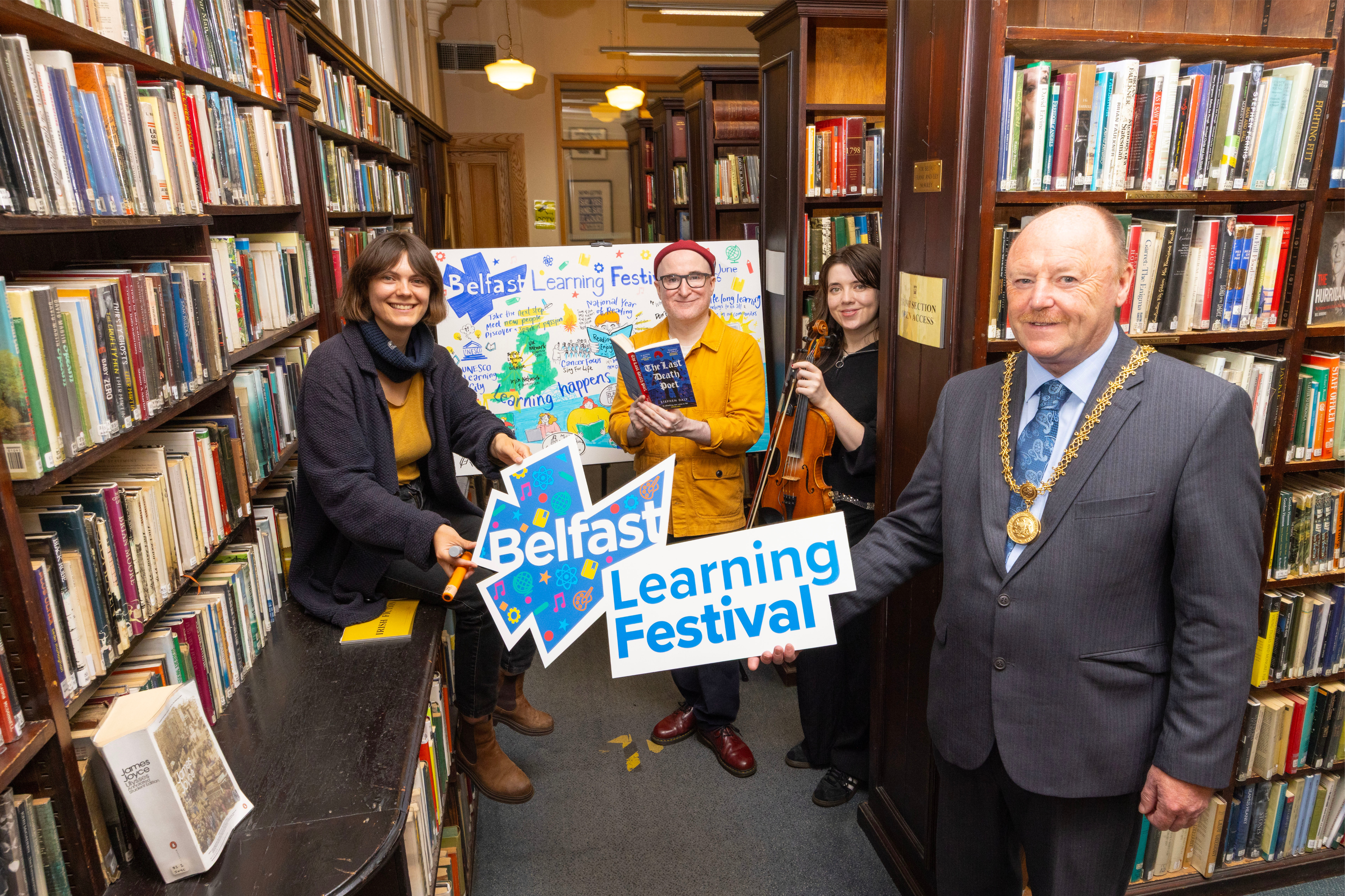 A group of people stand in a library.  One man holds a sign saying Belfast Learning Festival.  A lady sits in front of a large illustration, while a man holds a book and a young woman holds a fiddle.