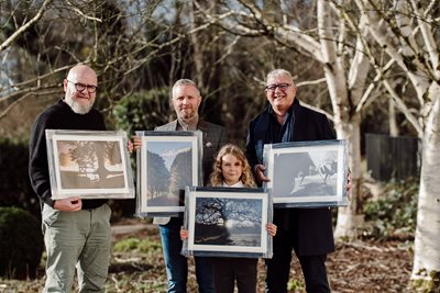 Four people, winners of the Belfast Parks Photographic Competition, hold winning framed photographs with trees behind them