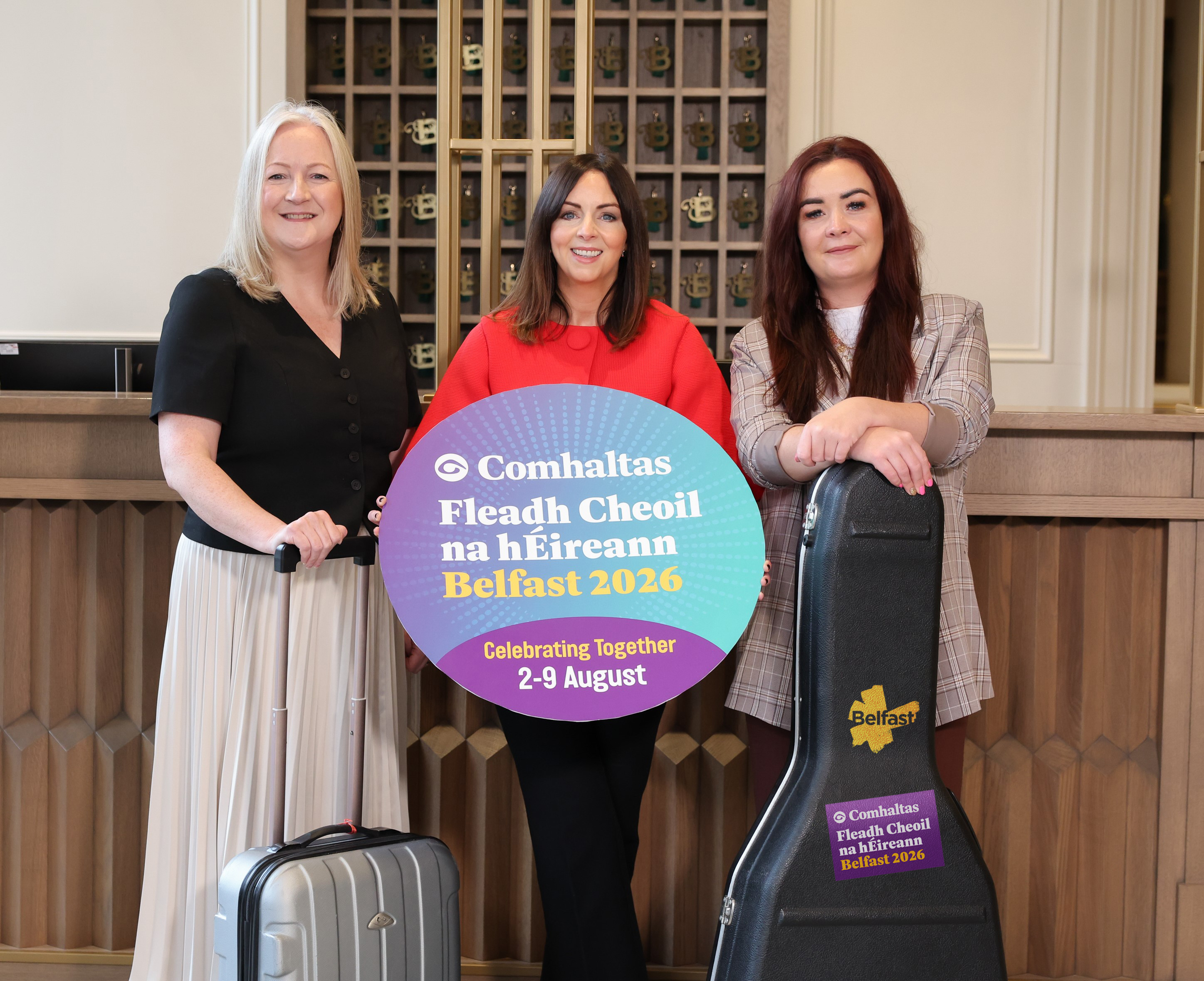 Three women gather at a hotel reception area, holding a suitcase, guitar case and holding a sign advertising Fleadh Cheoil na hÉireann.