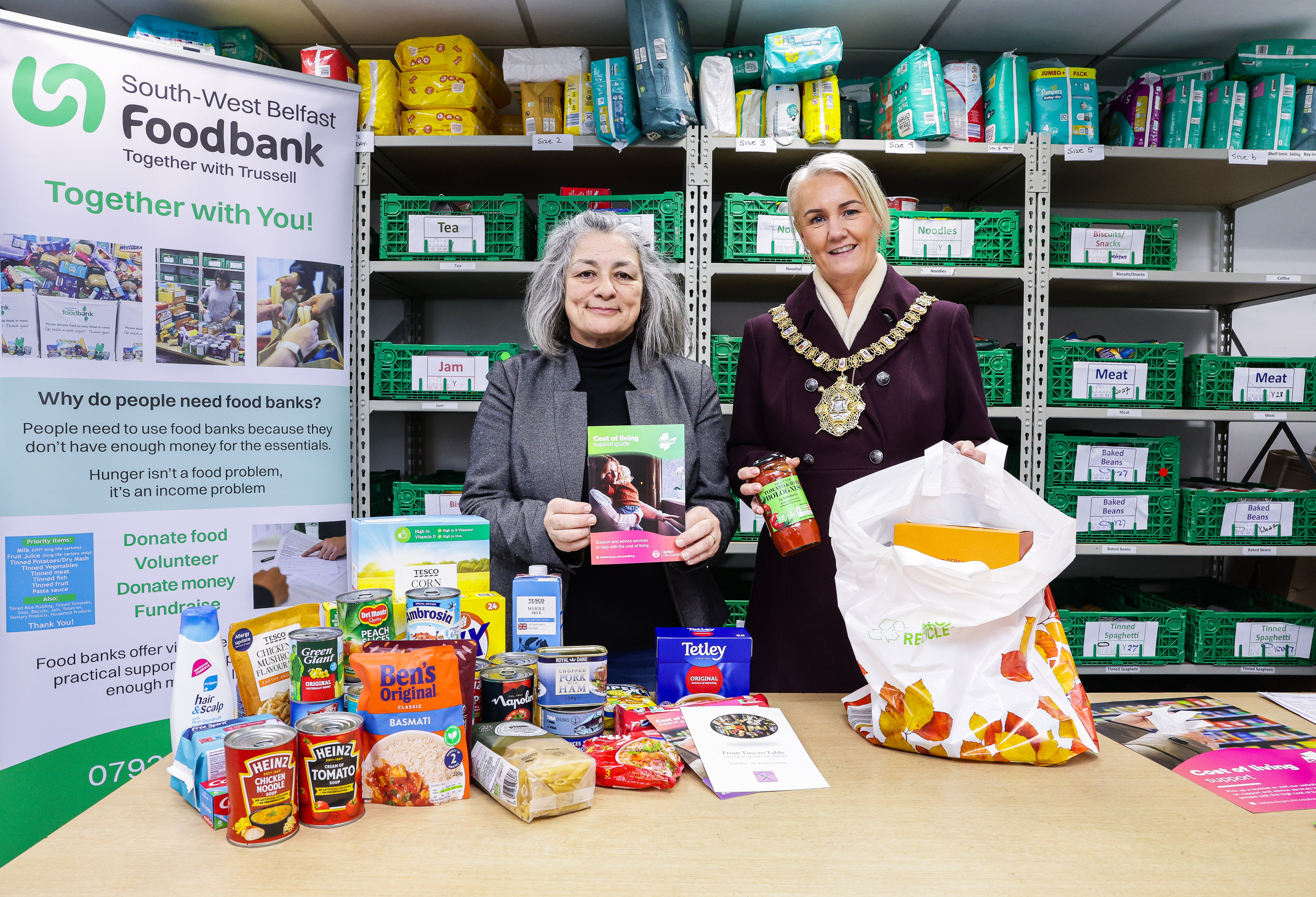 A rep from South West Belfast Foodbank with Lord Mayor Cllr Tracy Kelly in the foodbank with food items on a table and shelves in the background.