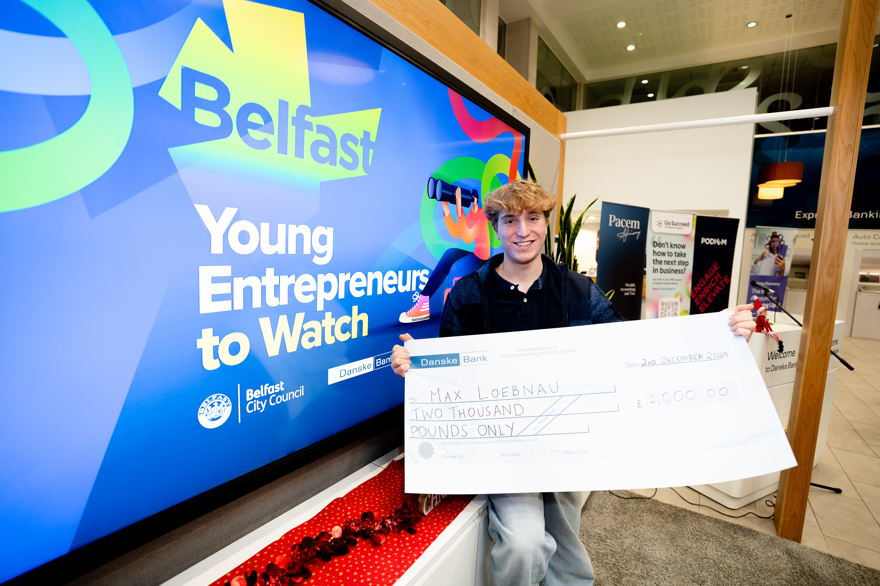 A young man holding a cheque for £2,000 in front of a screen displaying a colourful graphic which says 'Young Entrepreneurs to Watch'