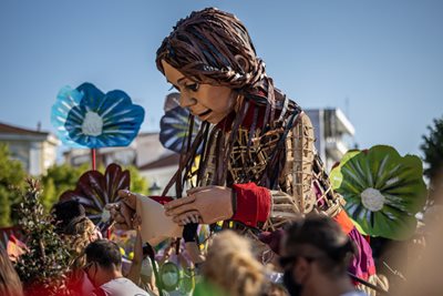 Little Amal, a giant puppet of a young girl, meets with visitors at an outdoor event.