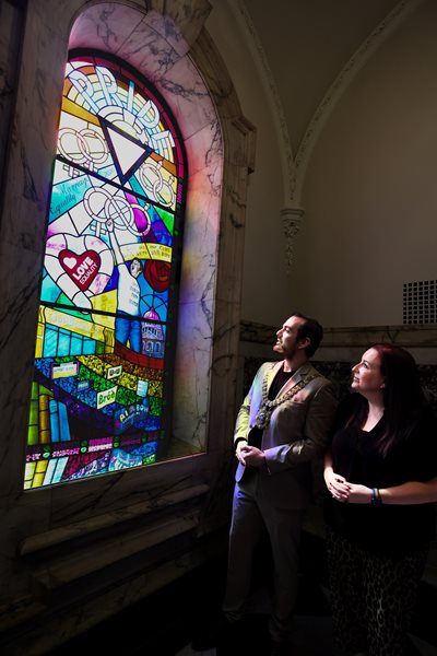 Lord Mayor of Belfast, Councillor Micky Murray, and Cara McCann, Director of HERe NI, beside a new stained glass window in City Hall, celebrating the contribution of the city’s LGBTQ+ community.