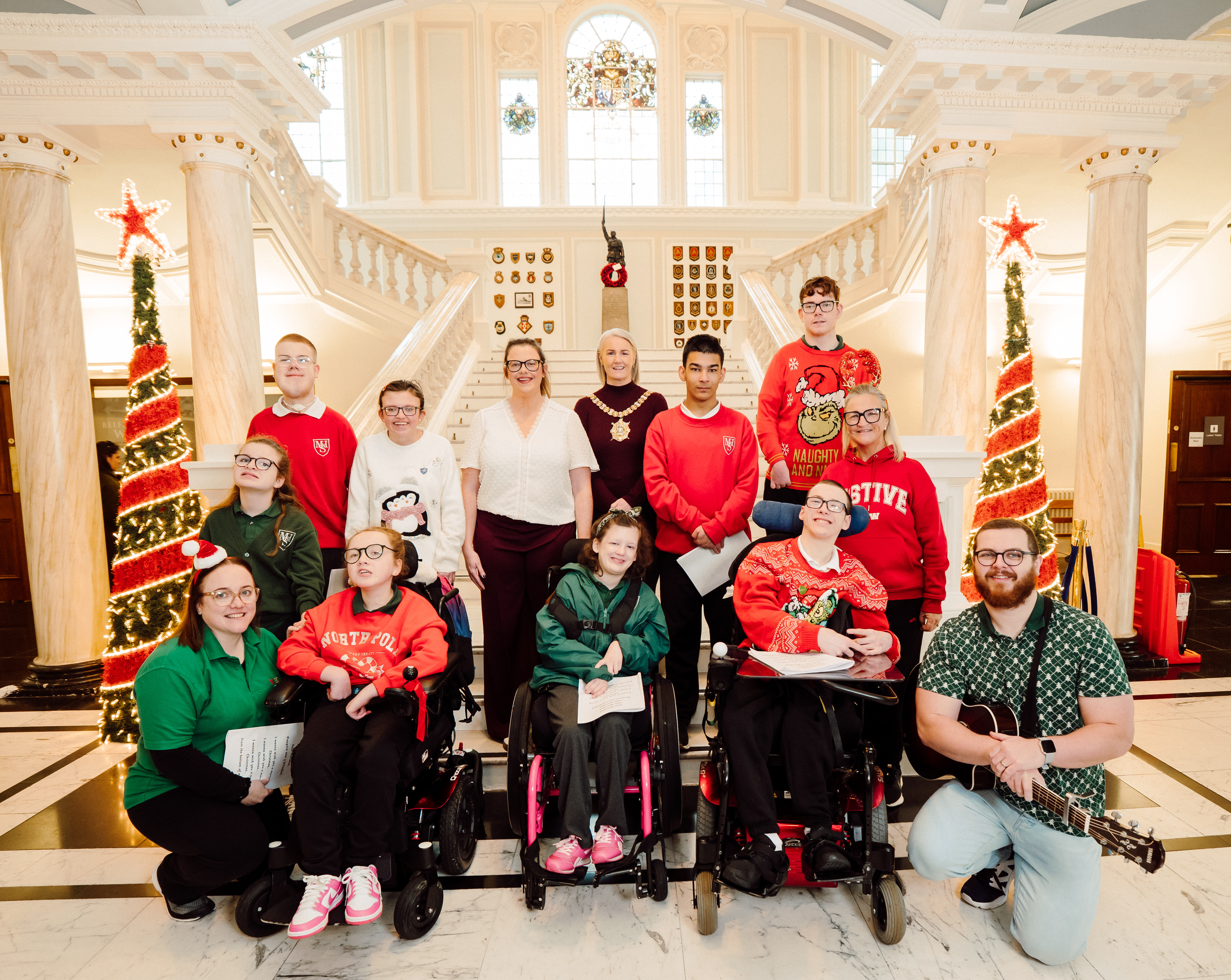 Lord Mayor Tracy Kelly, Councillor Ruth Brooks, Chair of the Disability Working Group and Mitchell House school choir in front of the staircase in City Hall with Christmas trees on either side.