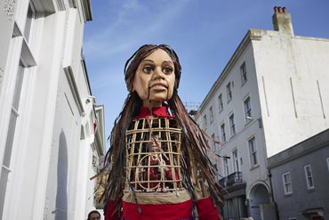 A photograph of the Little Amal puppet walking through an urban area