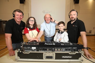 Councillor John Kyle wearing headphones, standing at a mixing desk. He is joined by two children and two men,