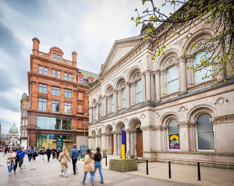 people walking past Grade B+ Listed Building 2 Royal Avenue in Belfast city centre, with Primark (Bank Buildings) next door and Belfast City Hall in the background.