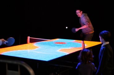 A colourful photograph of the artist playing ping-pong across a yellow and blue digital display table.