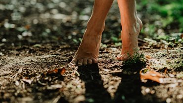 Photograph of a woman's bare ankles and feet walking across dry soil and grass