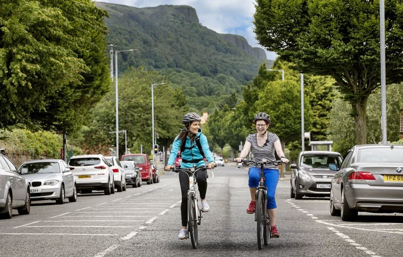 Two women cycling on Belfast’s Antrim Road, with Cave Hill in the background.