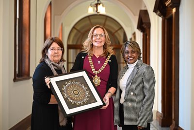 Lord Mayor Councillor Christina Black standing in a corridor of City Hall with Sheila Chakravarti and Ana Chandran from Belfast Asian Women’s Academy. They are holding a handmade mandala artwork.