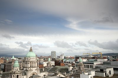 Belfast city skyline with Belfast City Hall dome visible against a cloudy blue sky.